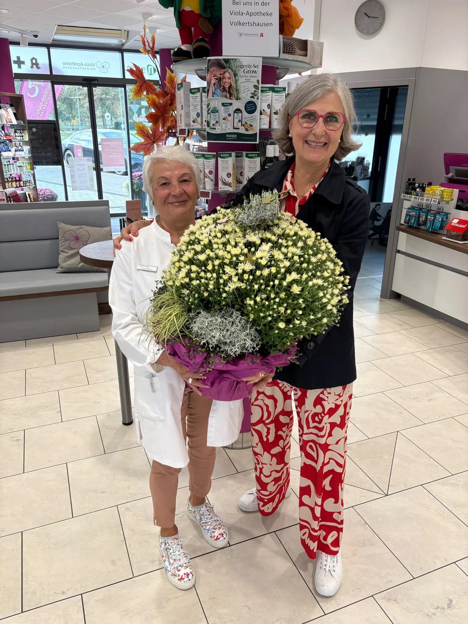 Rosanna Mancino im weißen Kittel und Apothekeninhaberin Stéphanie Viola Haas-Komp halten gemeinsam einen großen Blumentopf mit weißen Chrysanthemen in der Viola-Apotheke Volkertshausen