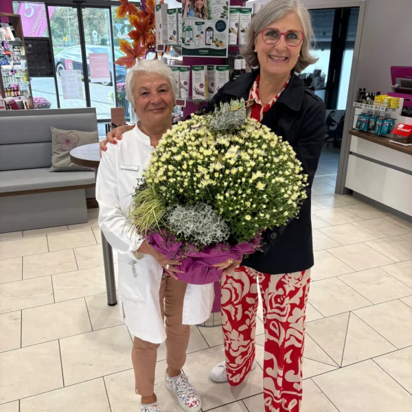 Rosanna Mancino im weißen Kittel und Apothekeninhaberin Stéphanie Viola Haas-Komp halten gemeinsam einen großen Blumentopf mit weißen Chrysanthemen in der Viola-Apotheke Volkertshausen