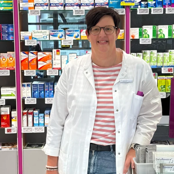 Marlene Wehrle im weißen Apothekerkittel und gestreiftem Shirt lächelt vor einem Regal mit Arzneimitteln in der Viola-Apotheke Volkertshausen