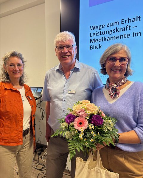 Apothekerin Stéphanie Viola Haas-Komp mit Blumenstrauß nach ihrem Vortrag beim VdK Ortsverband Oberer Hegau, neben ihr ein Mann und eine Frau vor der Präsentationsleinwand